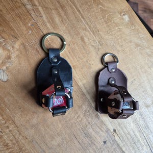 Può includere: Due porta dadi in pelle con portachiavi. Uno è nero con un anello dorato e un dado rosso visibile. L'altro è marrone con un anello argentato e un dado visibile. Entrambi sono su una superficie di legno.