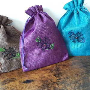May include: Three small drawstring bags made of burlap with a spiderweb design embroidered on the front. The bags are brown, purple, and teal.
