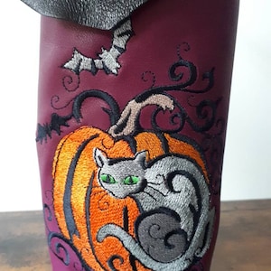 May include: A burgundy leather wine bottle holder with an embroidered Halloween design. The design features a gray cat with green eyes, an orange pumpkin, and a bat. The top of the holder is black.