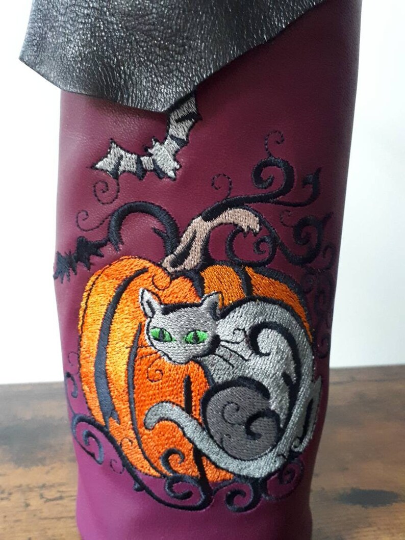 May include: A burgundy leather wine bottle holder with an embroidered Halloween design. The design features a gray cat with green eyes, an orange pumpkin, and a bat. The top of the holder is black.