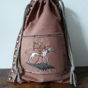 May include: A brown drawstring bag with a white rabbit riding a white dog embroidered on the front. The bag has silver metal studs around the edges and a green drawstring.