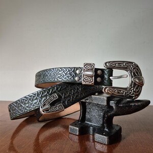 May include: A dark green leather belt featuring an embossed Celtic knot pattern throughout the strap. The belt includes a silver-toned metal buckle and a matching metal keeper, both decorated with intricate, raised knotwork designs.