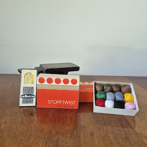 Op de afbeelding: Een verzameling naaibenodigdheden op een houten oppervlak. De items omvatten een doos met naalden, een doos met rood en wit garen met het opschrift "STOPFTWIST aus Viscose" en een doos met kleurrijke bollen wol. Een zwart object staat op de achtergrond.
