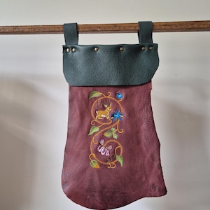 May include: A burgundy leather pouch with a dark green top and straps, adorned with an embroidered design of a rabbit, flowers, and vines. The pouch is suspended from a wooden measuring stick.