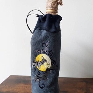 May include: A dark blue bottle cover with an embroidered design of a yellow moon and bats. The bottle has a gold cap, wrapped with twine, and a black handle. The design is suitable for Halloween.