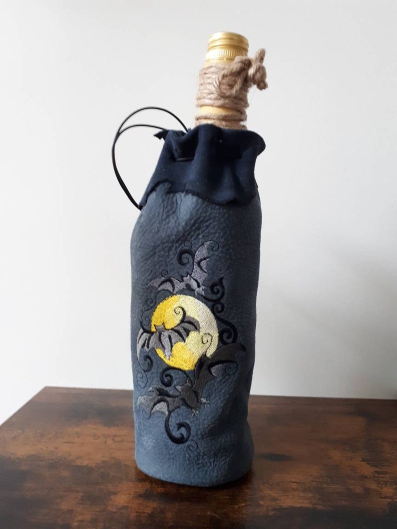 May include: A dark blue bottle cover with an embroidered design of a yellow moon and bats. The bottle has a gold cap, wrapped with twine, and a black handle. The design is suitable for Halloween.