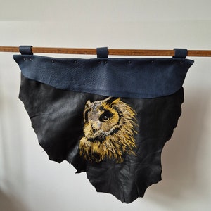 May include: A black leather wall hanging with a gold embroidered owl design. The hanging has a blue leather border and is attached to a wooden dowel.