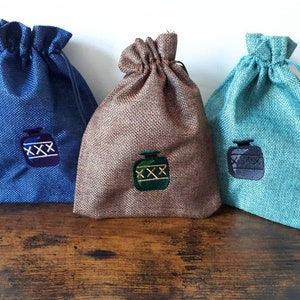 Könnte beinhalten: Drei Kordelbeutel aus Jutegewebe. Die Beutel sind in Blau, Braun und Türkis. Jeder Beutel hat ein gesticktes Design einer Flasche mit den Buchstaben 'XXX' darauf.