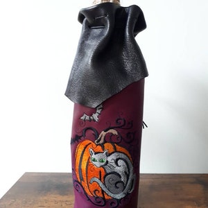 May include: A Halloween-themed wine bottle cover. The purple cover features an embroidered orange pumpkin with a gray cat and a bat. A black leather cape covers the top, and a rope secures the bottle.