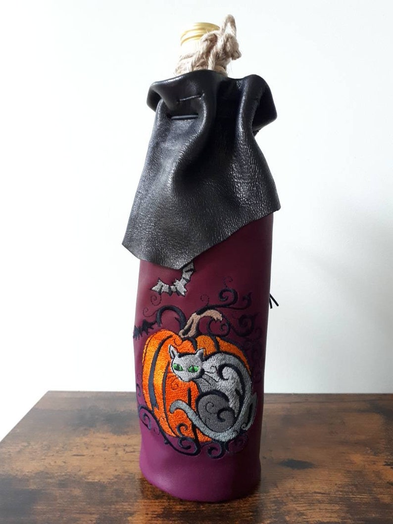 May include: A Halloween-themed wine bottle cover. The purple cover features an embroidered orange pumpkin with a gray cat and a bat. A black leather cape covers the top, and a rope secures the bottle.