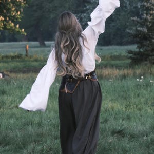 May include: A person in a white blouse with flared sleeves and dark pants, holding an object aloft in a grassy field. The outfit includes a brown belt with a pouch. The scene is set outdoors with trees in the background.