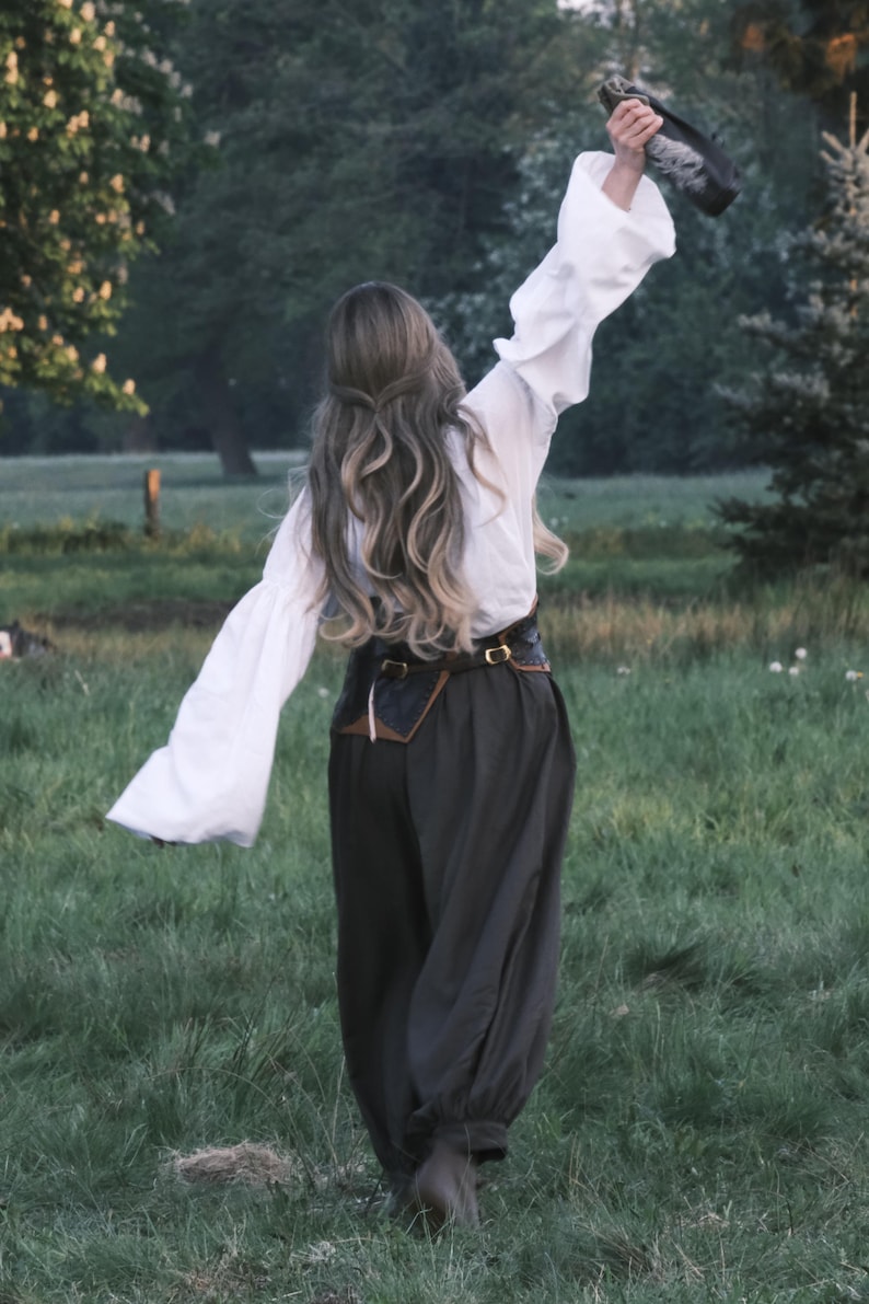 May include: A person in a white blouse with flared sleeves and dark pants, holding an object aloft in a grassy field. The outfit includes a brown belt with a pouch. The scene is set outdoors with trees in the background.