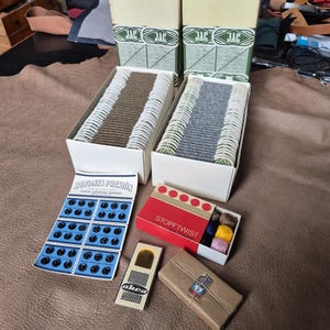 Puede incluir: Un surtido de suministros de costura, que incluye cajas de hilo Maaswol, cajas de hilo, una tarjeta de botones a presión negros y otros artículos de costura. Los artículos están dispuestos sobre una superficie de cuero marrón.