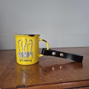 Könnte beinhalten: Gelbe Emaille-Tasse mit schwarzem Rand und Henkel. Die Tasse zeigt eine Grafik von Lemuren und den Text "gad company". Ein schwarzer Ledergriff ist mit zwei silbernen Nieten an der Tasse befestigt.