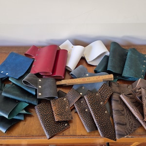 Puede incluir: Una colección de piezas de cuero en varios colores, incluyendo azul, verde, rojo y marrón. Las piezas están cortadas en diferentes formas y tamaños, y algunas tienen costuras o remaches decorativos. Las piezas están dispuestas sobre una superficie de madera.