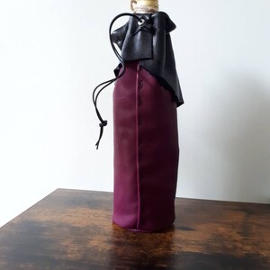 May include: A wine bottle sleeve made of deep purple leather, with a black leather top and drawstring closure. The bottle's gold-colored cap is visible at the top. The sleeve is designed to hold a standard-sized wine bottle.