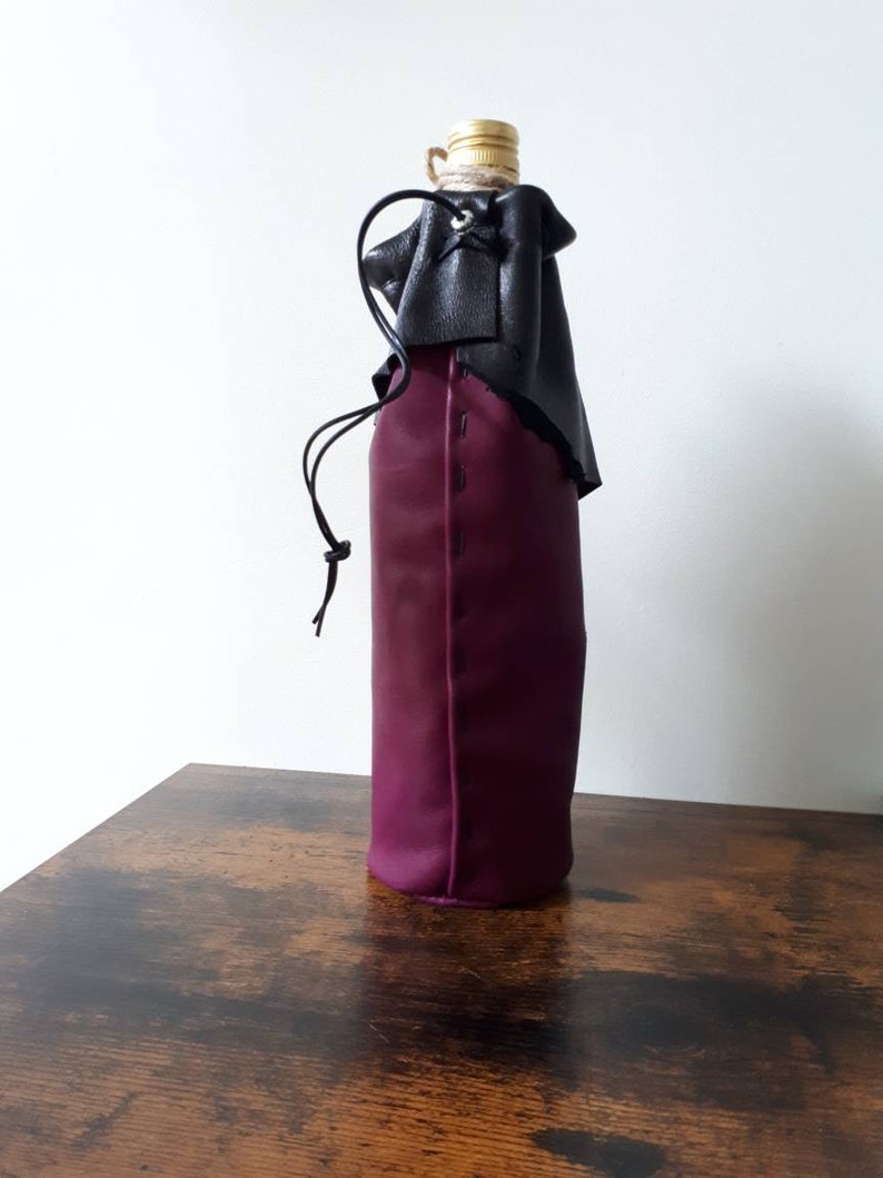 May include: A wine bottle sleeve made of deep purple leather, with a black leather top and drawstring closure. The bottle's gold-colored cap is visible at the top. The sleeve is designed to hold a standard-sized wine bottle.