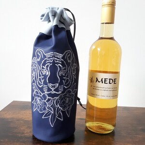 May include: A navy blue wine bottle bag with a white embroidered tiger face and floral design. A bottle of wine with a gold cap and a label with text is next to the bag. The bag has a drawstring closure.
