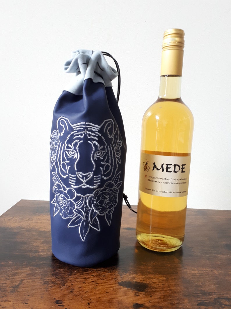 May include: A navy blue wine bottle bag with a white embroidered tiger face and floral design. A bottle of wine with a gold cap and a label with text is next to the bag. The bag has a drawstring closure.