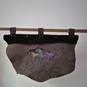 May include: A grey leather wall hanging featuring an embroidered design of three galloping horses in tan, blue, and purple. The piece has a black leather top border secured with metal rivets and three dark leather hanging loops on a wooden rod.