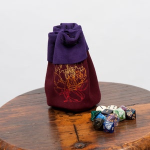 Può includere: Un sacchetto da gioco in tessuto viola e bordeaux con un disegno ricamato in oro. Il sacchetto è appoggiato su un tavolo di legno con un set di dadi colorati sparsi intorno.