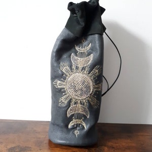 May include: A dark gray leather wine bottle bag with a black drawstring closure. The bag is adorned with an elaborate sunburst and crescent moon design in gold-colored beads. The bag is designed to hold a standard-sized wine bottle.