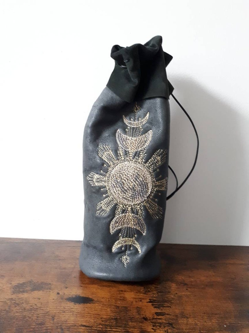 May include: A dark gray leather wine bottle bag with a black drawstring closure. The bag is adorned with an elaborate sunburst and crescent moon design in gold-colored beads. The bag is designed to hold a standard-sized wine bottle.