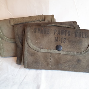 Puede incluir: Tres bolsas de lona color verde oliva, una de ellas con la etiqueta "Spare Parts Roll M-13".