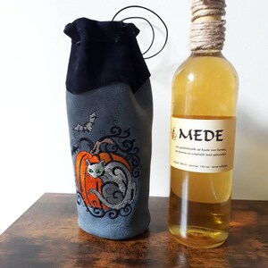 May include: A gray wine bottle bag with an embroidered orange pumpkin and a gray cat design. A bat is embroidered above the pumpkin. A bottle of mead with a rope-wrapped neck is next to the bag.