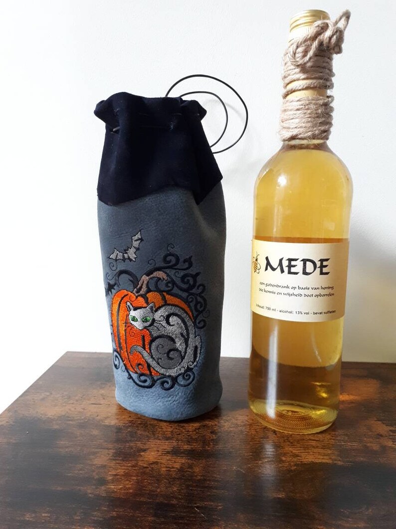 May include: A gray wine bottle bag with an embroidered orange pumpkin and a gray cat design. A bat is embroidered above the pumpkin. A bottle of mead with a rope-wrapped neck is next to the bag.