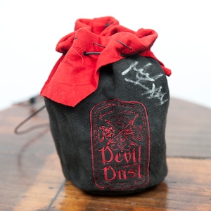 Może przedstawiać: Czarna torba ze sznurkiem do ściągania z czerwoną klapą i haftowanym czerwonym logo z napisem "Devil Dust".