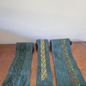 Puede incluir: Tres rollos de tela verde azulado con diseños bordados. Uno presenta un patrón negro ornamentado, otro tiene un diseño floral colorido y el tercero muestra un borde floral rojo y verde. La tela está sobre una superficie de madera.