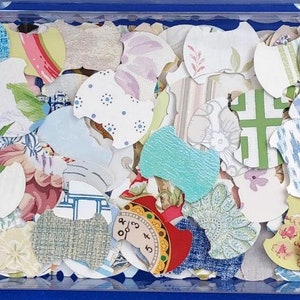 May include: A clear plastic container filled with a variety of colorful and patterned paper scraps. The scraps are cut into different shapes, including circles, squares, and rectangles. Some of the scraps have floral designs, while others have geometric patterns.
