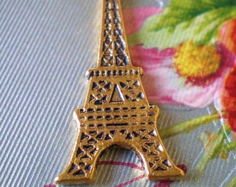 May include: A gold-coloured Eiffel Tower charm with detailed features. The charm has a loop at the top for attachment and features dark blue accents within the tower's design. The charm is set against a light blue background with floral accents.