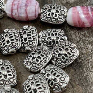 May include: A close-up of silver metal beads with a decorative floral design. The beads are square shaped and have a raised design.