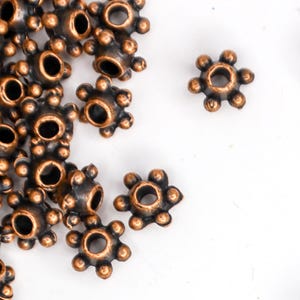 May include: A pile of copper-colored metal beads with a starburst design. Each bead has a small hole in the center for stringing.