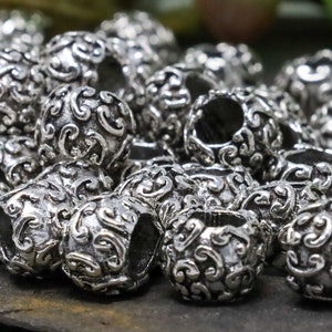May include: A collection of silver-colored, round beads with an ornate, swirling pattern. The beads have a hollow center and are clustered together, showcasing their detailed design. The background features blurred green foliage.