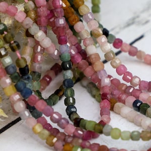 May include: A strand of multi-colored gemstone beads. The beads are small and square-shaped. The colors include pink, green, blue, and brown.