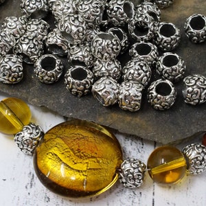 May include: Silver-colored metal beads with an ornate, swirling design are scattered across a dark surface. A necklace with large, amber-colored glass beads and matching silver beads is visible at the bottom. The beads are likely for jewelry making.