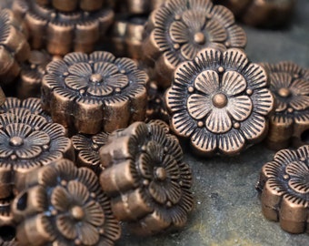 Può includere: Un mucchio di perline di metallo color rame a forma di fiore con un foro al centro di ogni perlina. Le perline sono disposte in modo casuale.