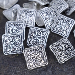 May include: A collection of silver-toned square buttons with intricate, ornate designs. The buttons have a raised, textured surface and a slightly rounded edge.