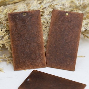 May include: Three rectangular tags with a distressed brown finish and a smooth, flat surface. Each tag features a single small circular hole punched near the top edge, designed for attachment. The tags have a weathered, rustic appearance.