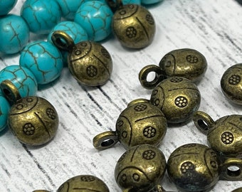 May include: A collection of antique gold-toned ladybug charms with intricate details. The charms are scattered on a white surface with a turquoise blue beaded necklace in the background.