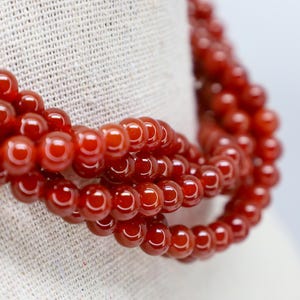 May include: A close-up of a necklace with multiple strands of glossy, reddish-orange beads. The beads are round and evenly sized, creating a smooth, flowing appearance. The necklace is draped over a light-colored fabric.
