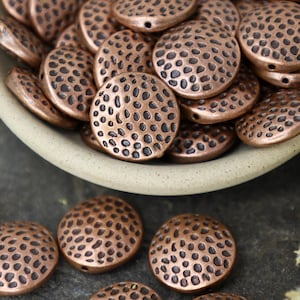 May include: A bowl filled with copper-colored round beads with a textured surface. The beads have a hammered or dimpled look. The bowl is sitting on a dark brown surface.