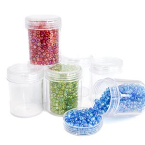 Puede incluir: Varios botes de plástico transparente llenos de cuentas de colores en tonos rojos, verdes y azules. Un bote está volcado, derramando cuentas azules. Los botes están dispuestos sobre una superficie blanca. Ideal para manualidades.