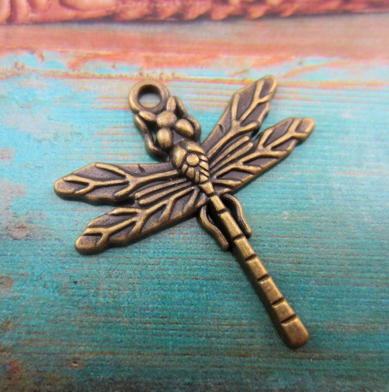 May include: Bronze-colored dragonfly charm with detailed wings and body. The charm has a loop at the top for attaching to jewelry or other crafts. The dragonfly is a popular symbol in jewelry and craft making.