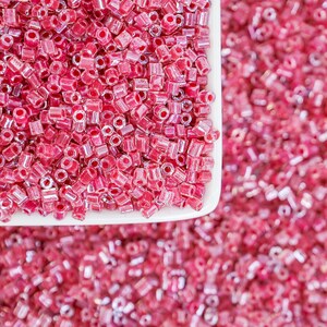 May include: A close-up shot of a white square dish filled with translucent, pink seed beads. The beads are scattered, with some spilling out onto a pink surface. The beads are small, cylindrical, and have a shiny, iridescent quality, perfect for jewelry making.