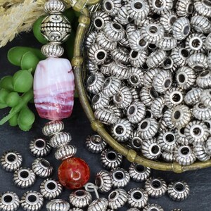 May include: A collection of small, round, silver-toned metal beads with a ribbed texture, alongside a single smooth red stone bead and a rectangular white and pink marbled glass bead, arranged with decorative silver-toned spacer beads.