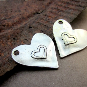 May include: Two silver heart-shaped charms with a smaller heart design in the center. The charms have a hole at the top for attaching to jewelry or other crafts.